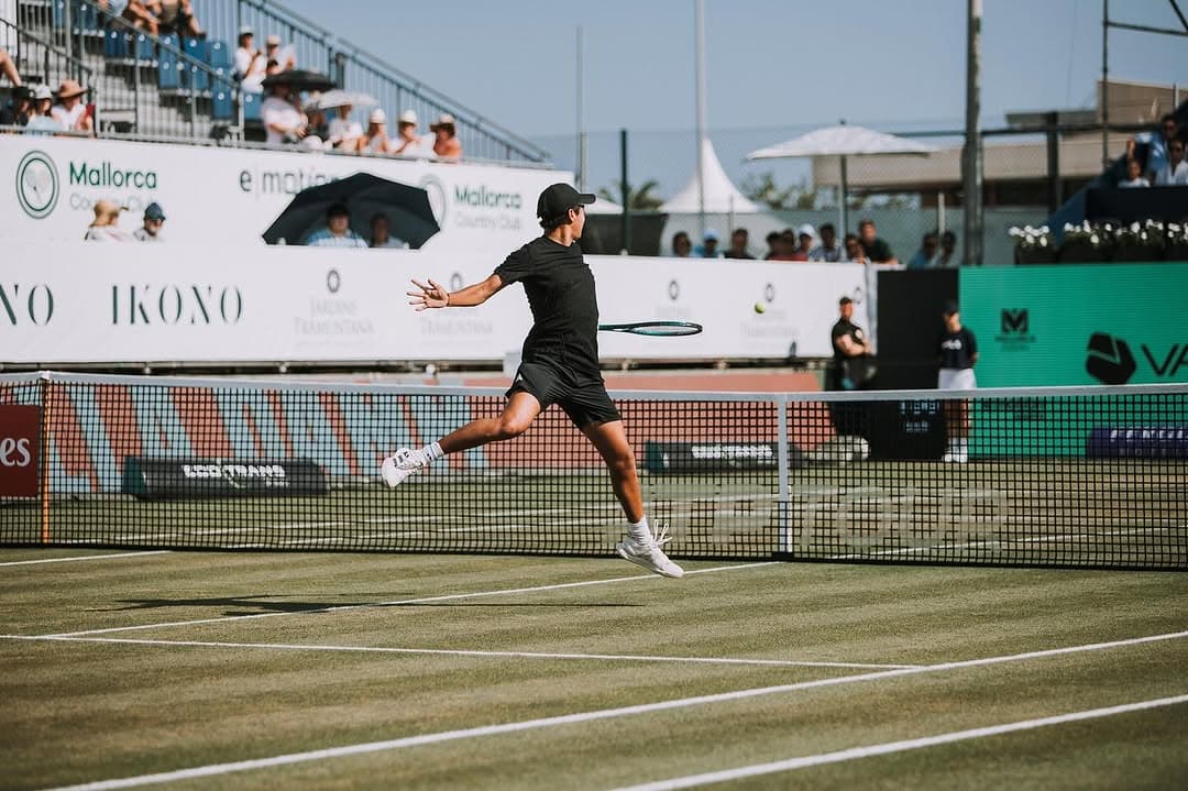 Learner follows through on a forehand on grass court, Mallorca and IKONO sponsors visible.