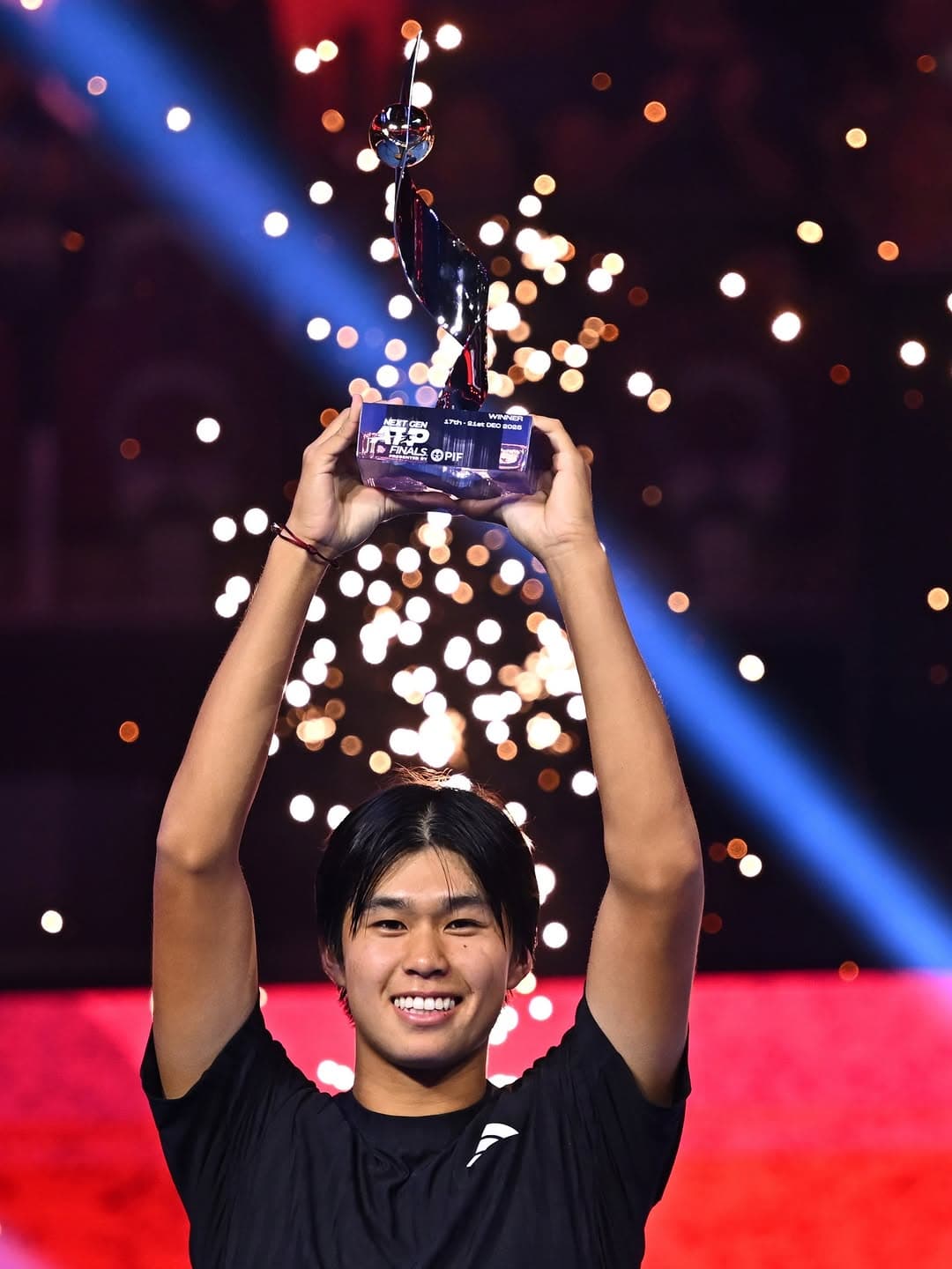 Learner Tien holds an ATP Finals trophy above his head, smiling on stage with colorful stage lights behind him.