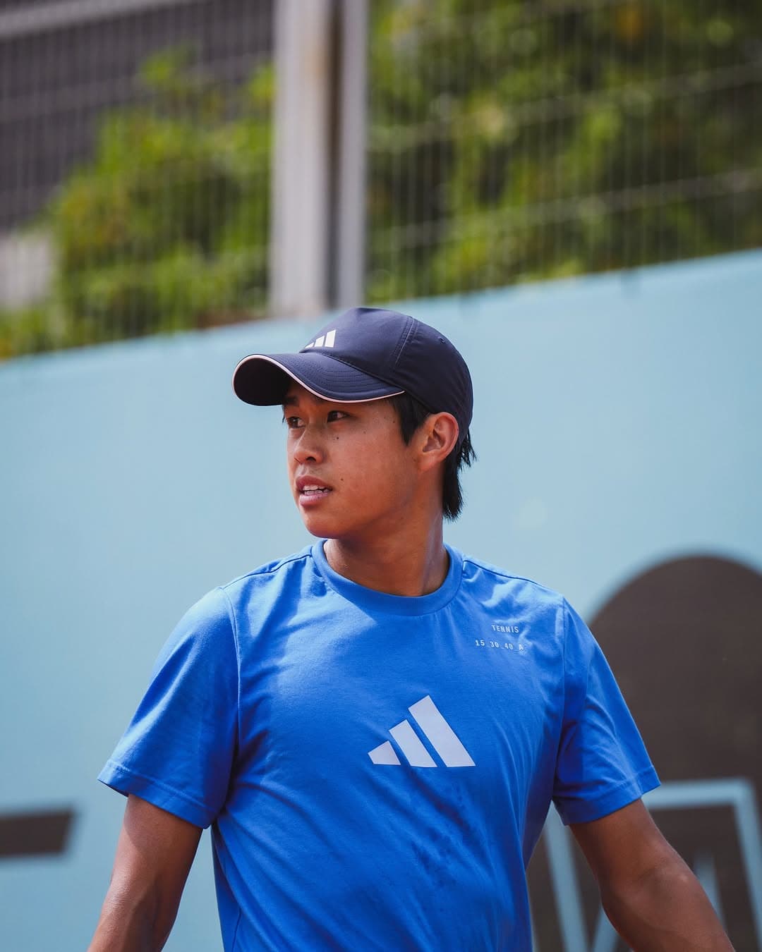 Learner Tien in a blue Adidas kit and navy cap, looking across the court between points.