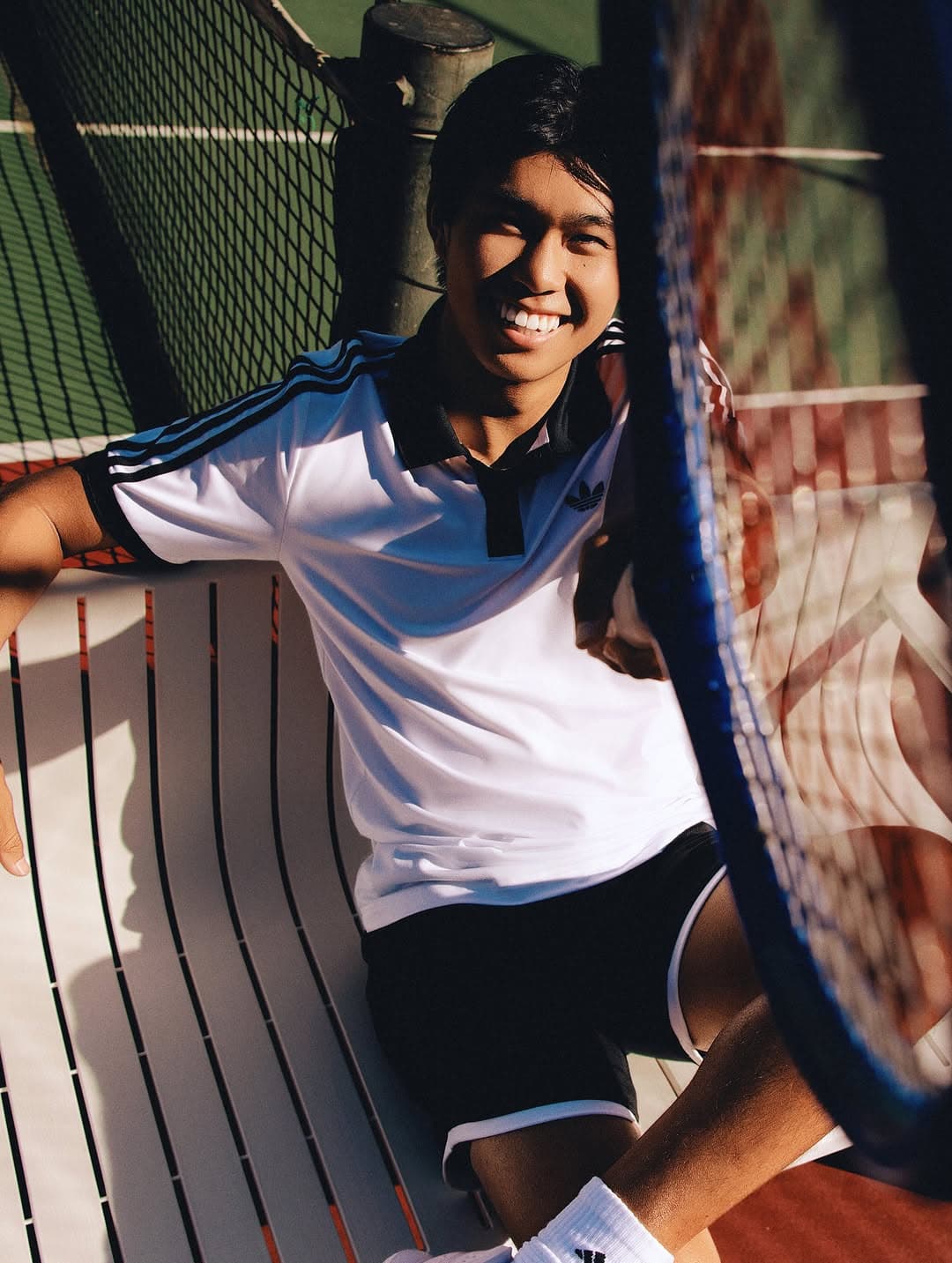 Learner smiles at the net on a clay court in bright sunlight, wearing blue-striped white tennis top.